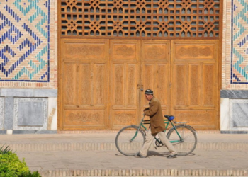 Homme en tenue ouzbèke tirant son vélo devant une célèbre mosquée en Ouzbékistan