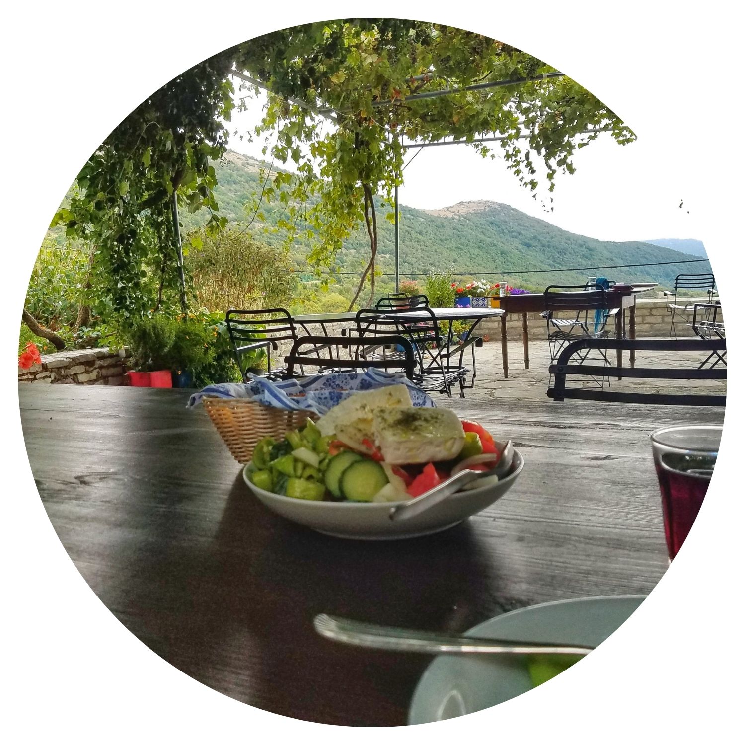Une salade grecque typique servie sur une terrasse offrant une vue pittoresque sur la nature verdoyante et les petites montagnes, ombragée par des feuilles d'arbre.