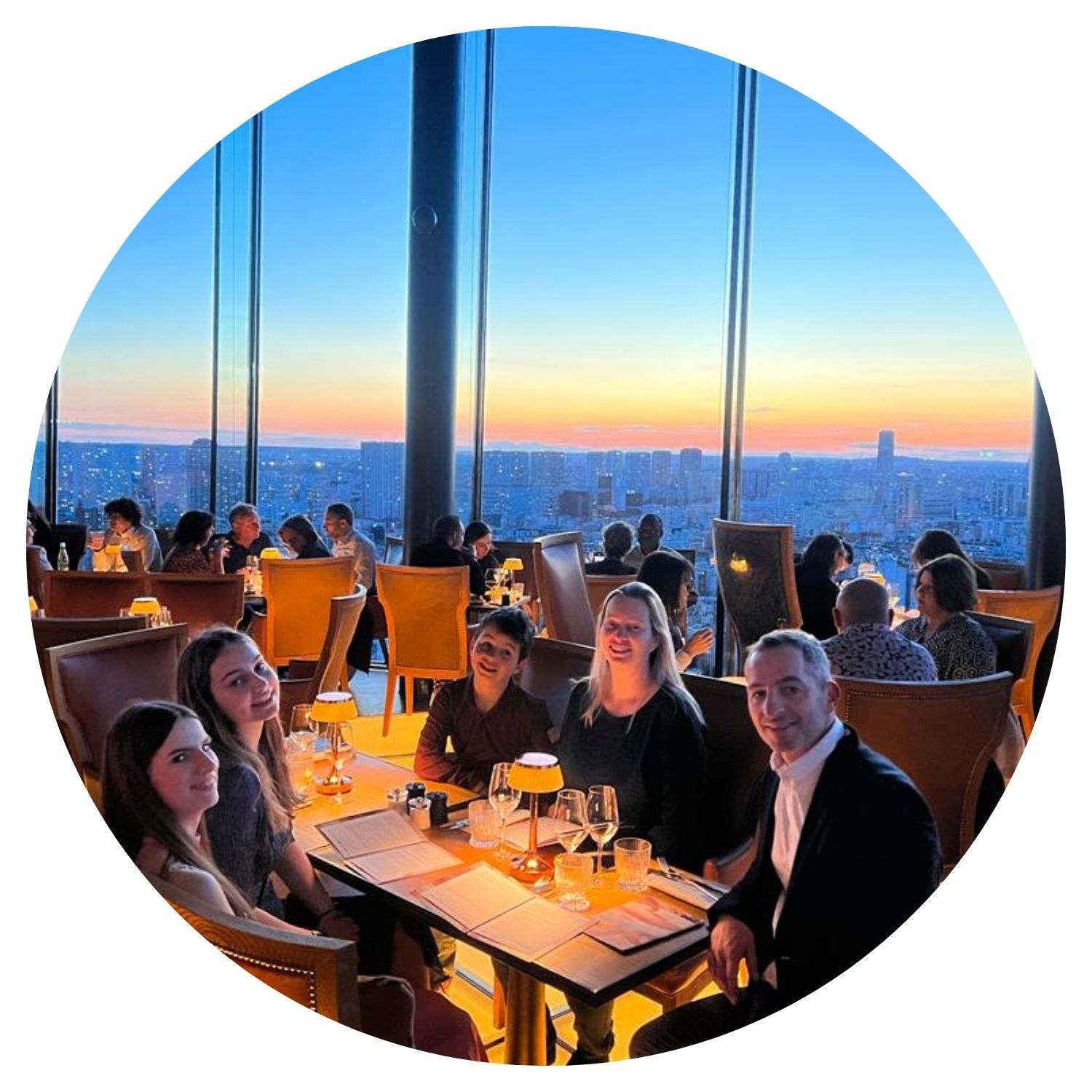 Une famille de cinq personnes dîne dans un restaurant élégant avec de grandes fenêtres offrant une vue panoramique sur New York. Les membres de la famille sont souriants et apprécient leur repas ensemble.