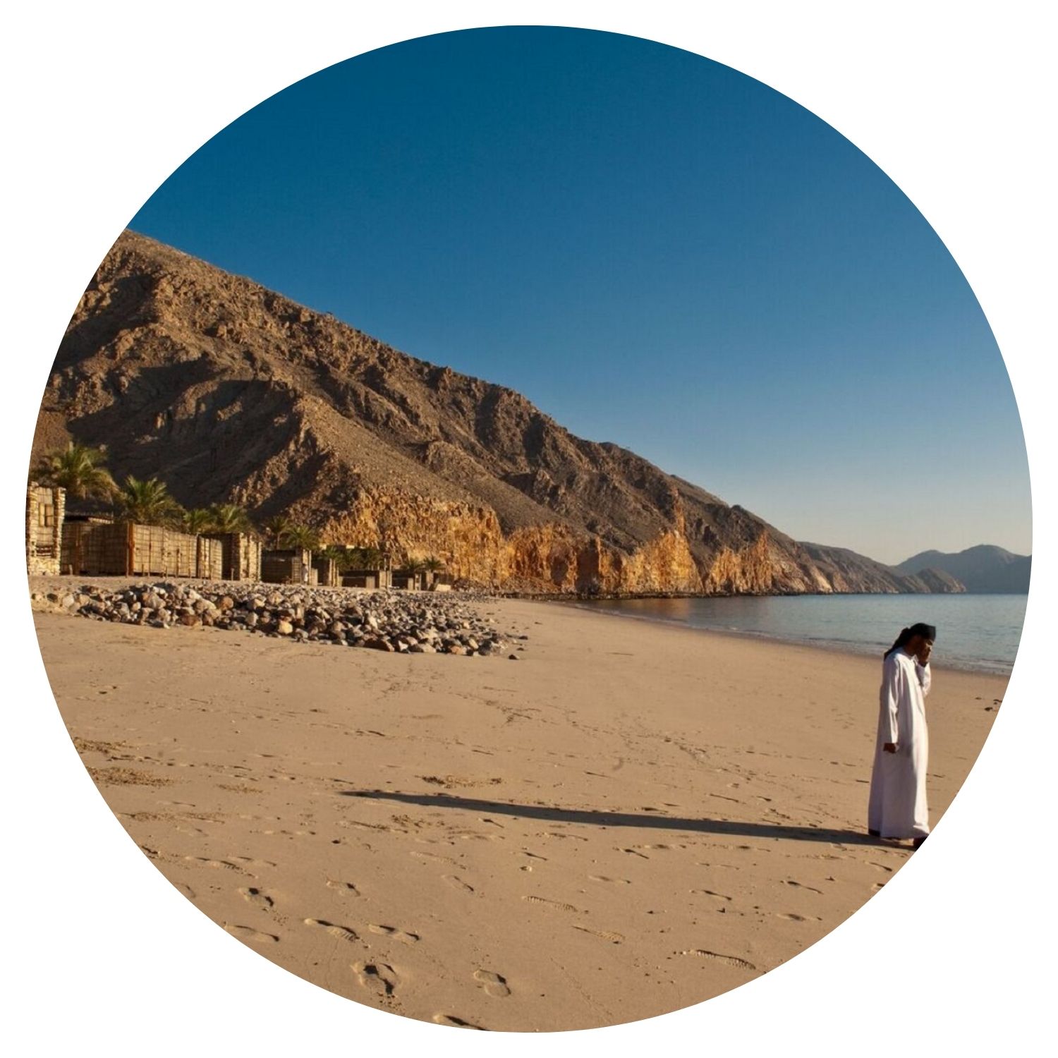 Un jeune homme, vêtu de la tenue traditionnelle omanaise, se tient debout sur une plage entourée de nature sauvage. La scène se déroule en Oman, offrant un aperçu authentique de la culture et des paysages naturels du pays.